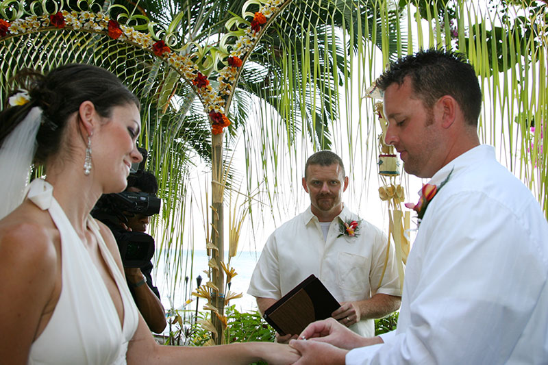 Couple exchanging rings under a palm-leaf and orchid arch with the celebrant behind them at Santai Hotel Amed in east Bali