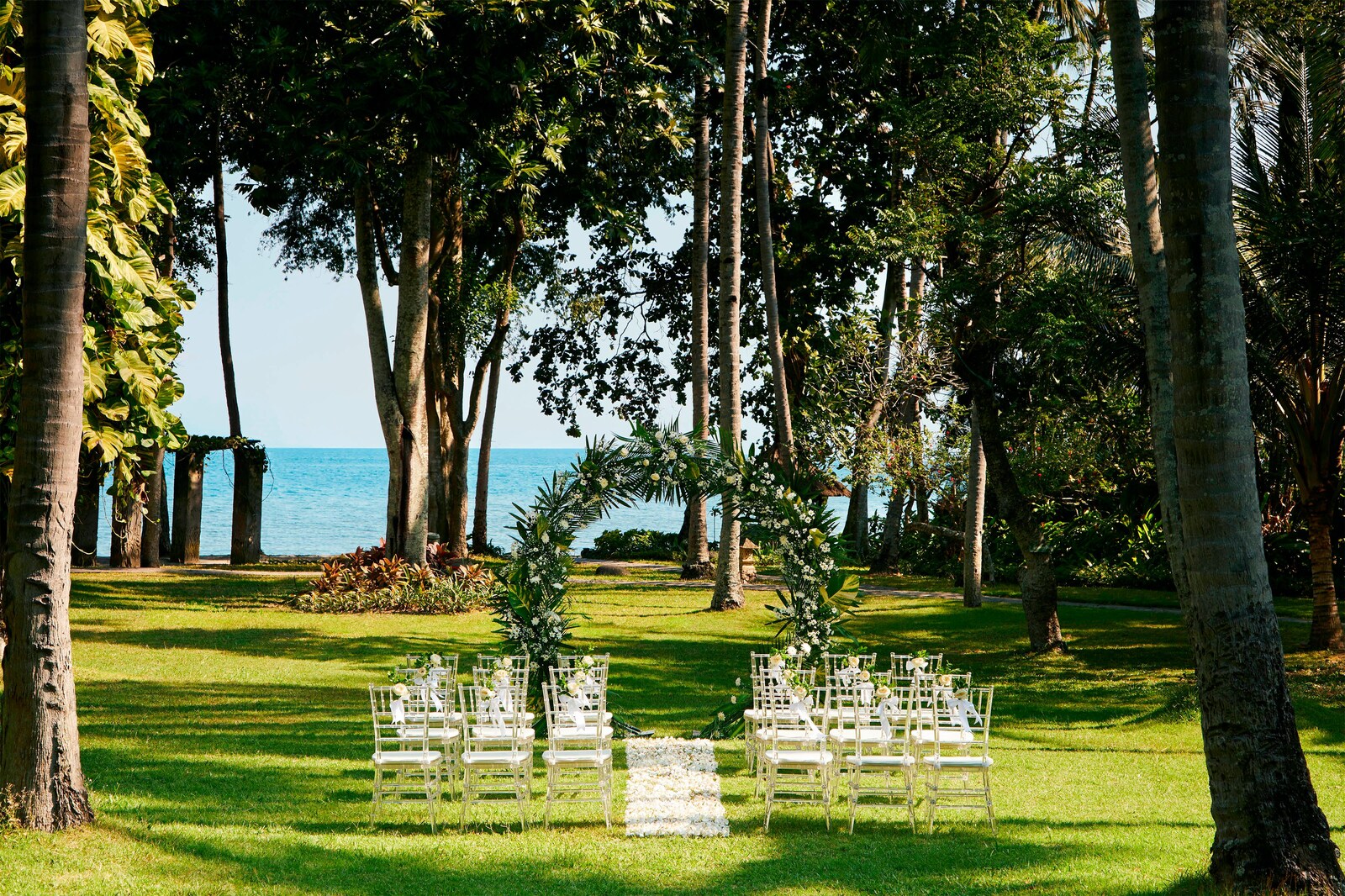 Ceremony chairs and a floral arch on the palm-shaded garden lawn facing the sea at Sheraton Senggigi Beach Resort, Lombok