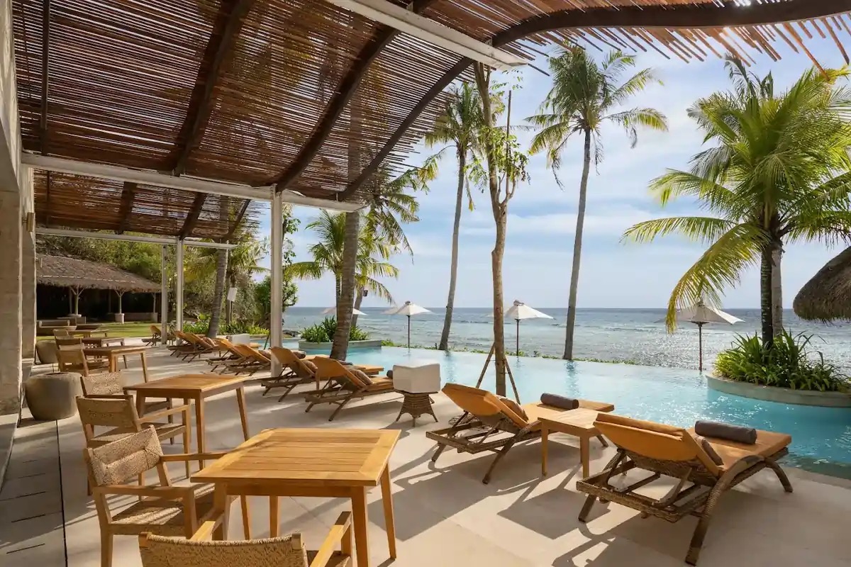 Shaded terrace with teak tables beside the pool and palms at Silo Beach Club on Nusa Penida, Bali