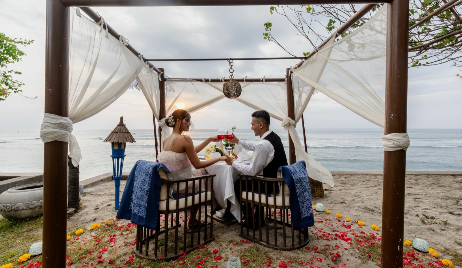 Couple toasting at a draped beachfront pergola at sunset with petals on the sand, Sudamala Resort, Senggigi