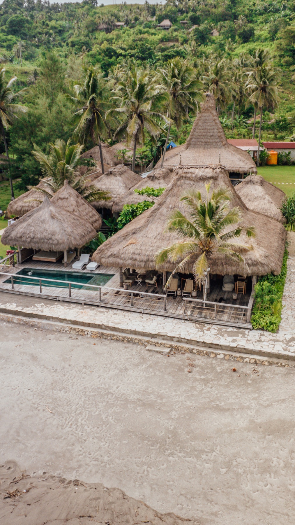 Aerial view of the thatched roofs and pool of Sumba Beach House above the beach, Sumba