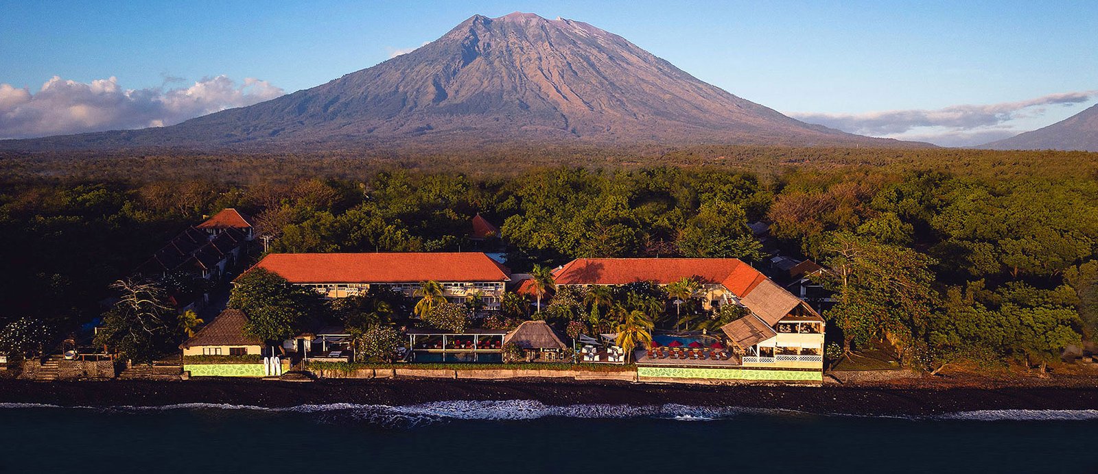 Aerial view of Tauch Terminal Resort Tulamben & Spa on the black sand shore with Mount Agung rising behind it in northeast Bali