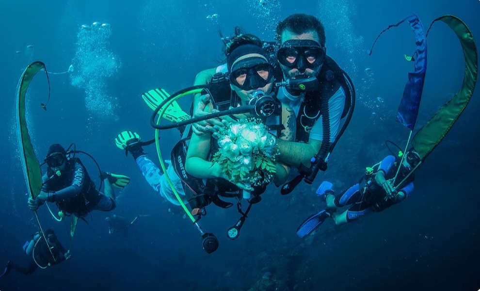 Couple diving together with a bouquet, escorted by divers holding ceremonial banners, during an underwater wedding at Tauch Terminal Resort Tulamben & Spa in northeast Bali
