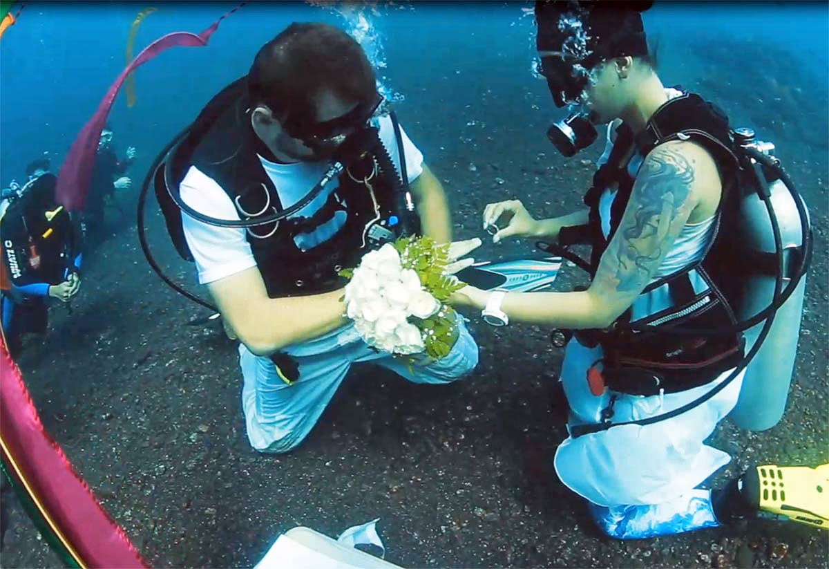 Bride and groom in scuba gear exchanging rings on the black sand seabed during an underwater ceremony at Tauch Terminal Resort Tulamben & Spa in northeast Bali