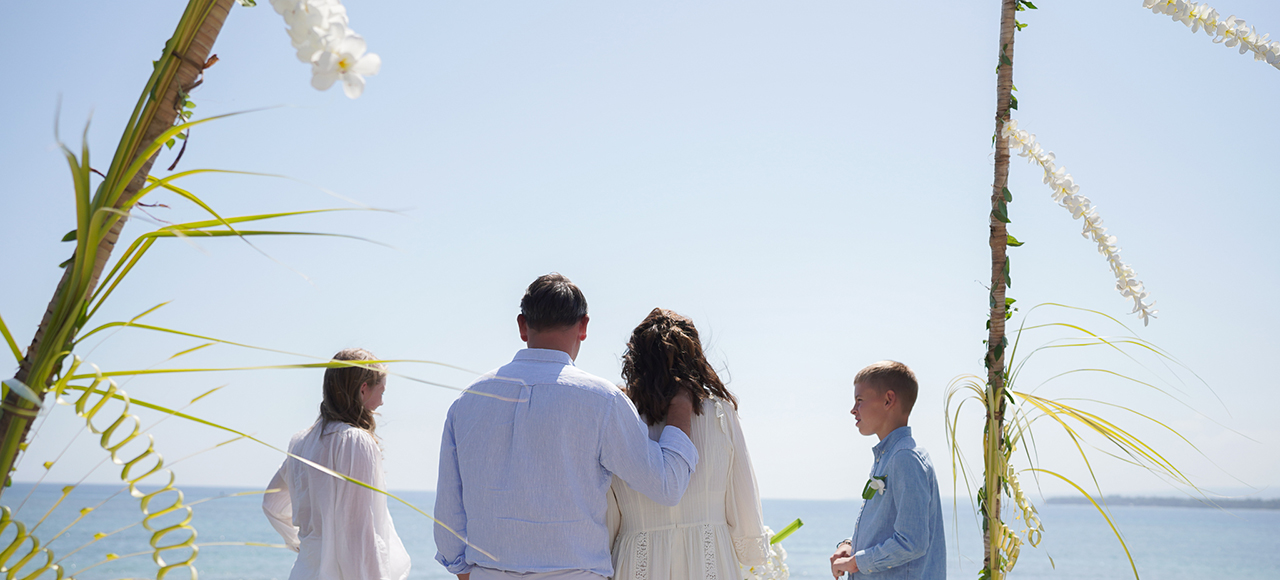 Small family group at a ceremony framed by frangipani-wrapped poles above the water at The Lombok Lodge, Medana, Lombok