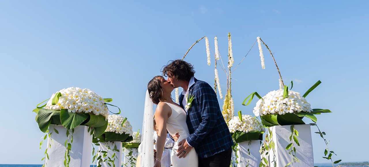 Couple kissing between pedestals of white frangipani under hanging flower strands at their ceremony at The Lombok Lodge, Medana Bay, Lombok