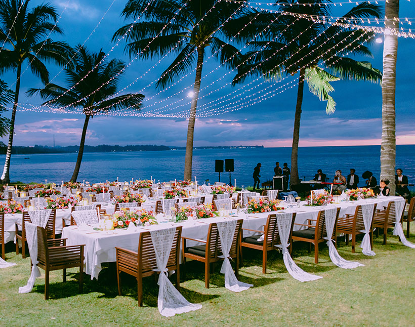 Long banquet tables under strings of lights among coconut palms at dusk, The Oberoi Beach Resort Lombok