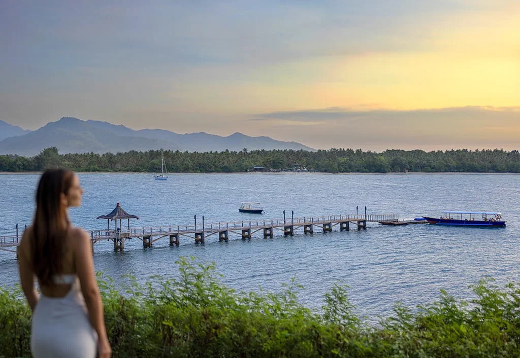 Woman in white looking out to the long wooden jetty at sunset at Medana Beach, The Oberoi Beach Resort Lombok