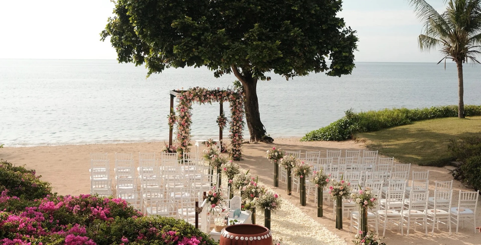 Ceremony set with a pink and peach floral arch under a spreading tree and rows of white chairs on the sand at The Oberoi Beach Resort Lombok, on Lombok's northwest coast
