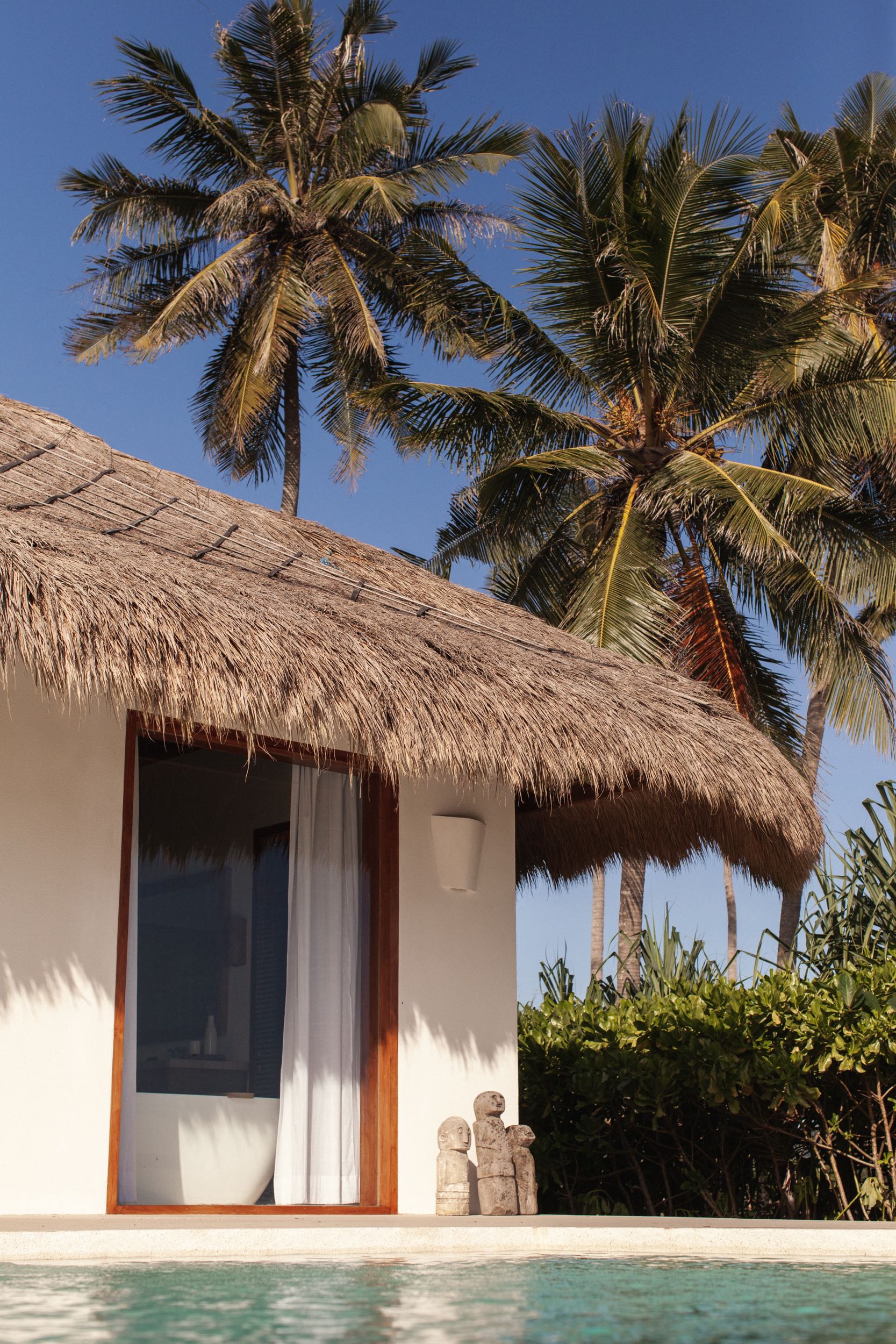 Thatched villa roof and private pool framed by coconut palms, with carved stone figures at the water's edge, at The Sanubari, Sumba