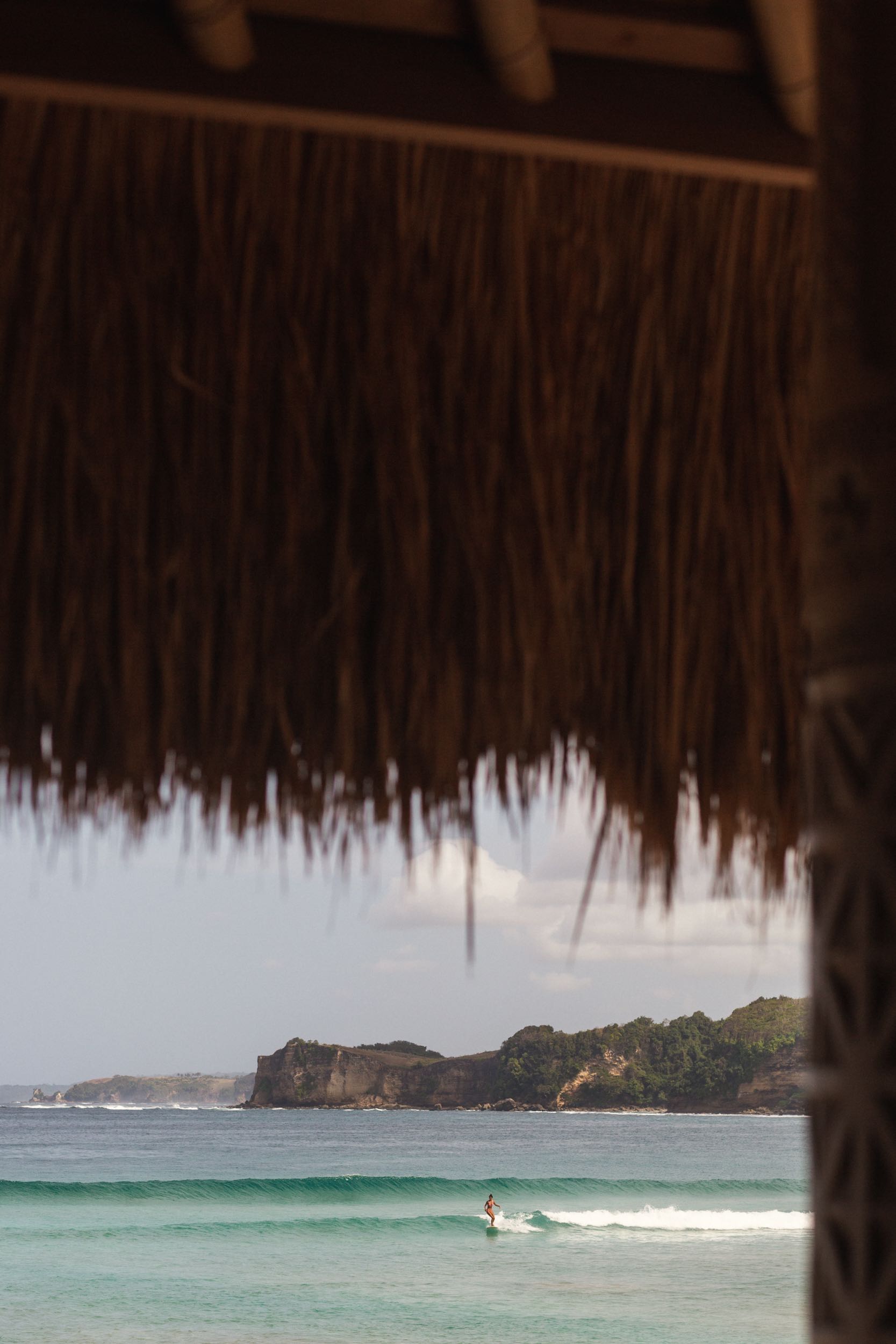 A surfer on a wave with the green headland behind, seen from under a thatched roof at The Sanubari, Sumba