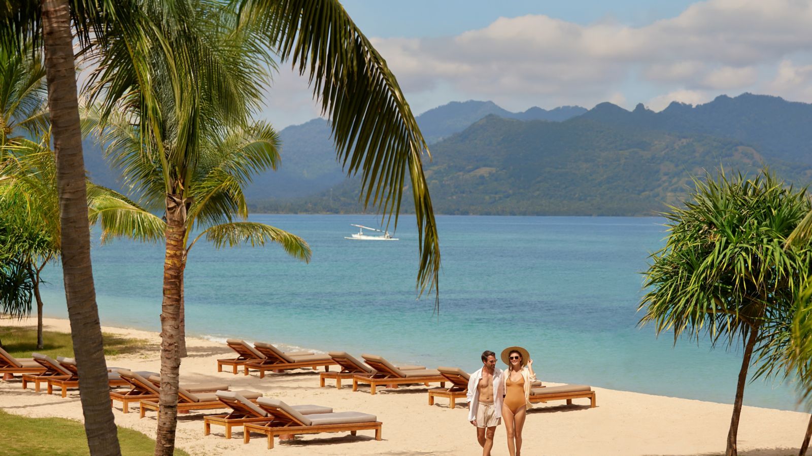 Sun loungers under palms on the beach at The Sira, with green hills across the water, Lombok