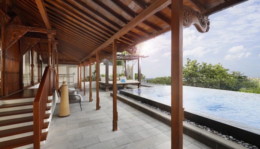 The carved timber terrace and infinity pool of The Tulou Bali on the cliff above Jimbaran, with the sea in the distance
