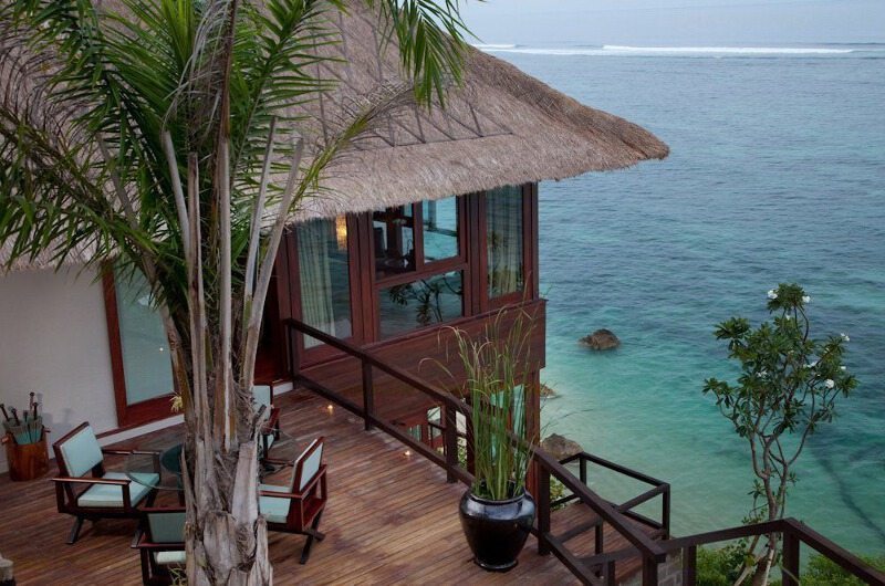 A thatched cliff-edge bale and deck at Villa Bidadari Cliffside Estate above the Indian Ocean, with surf breaking below and palms framing the view