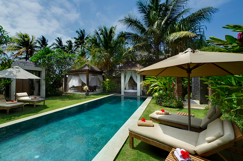 Pool bordered by lawn, palms, and thatched garden pavilions at Villa Maya, Majapahit Beach Villas