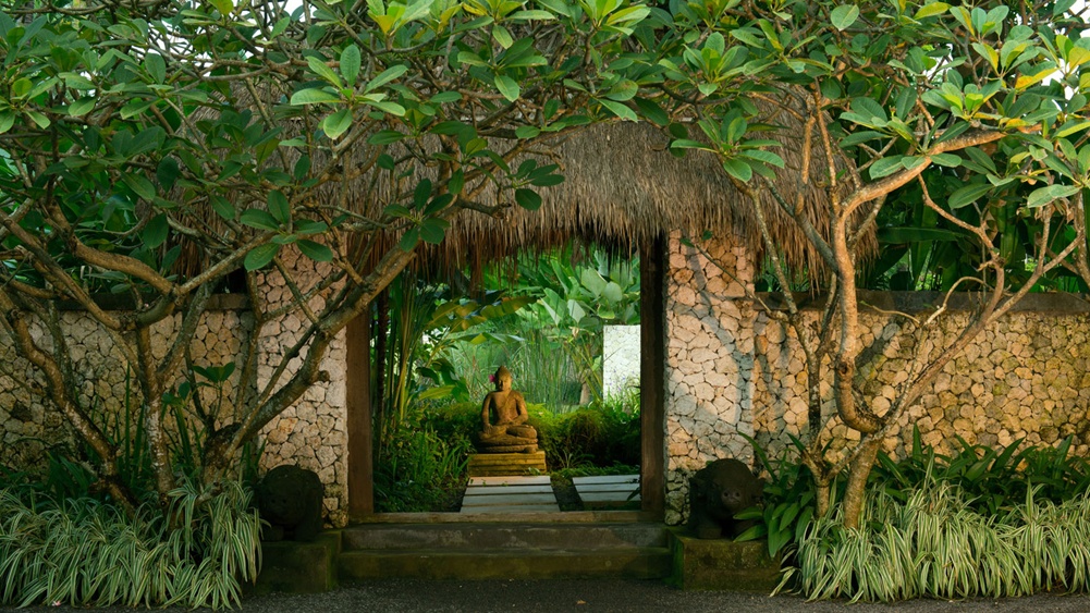 A stone garden gateway with a thatched roof, framing a seated stone Buddha in the greenery at Villa Samadhana