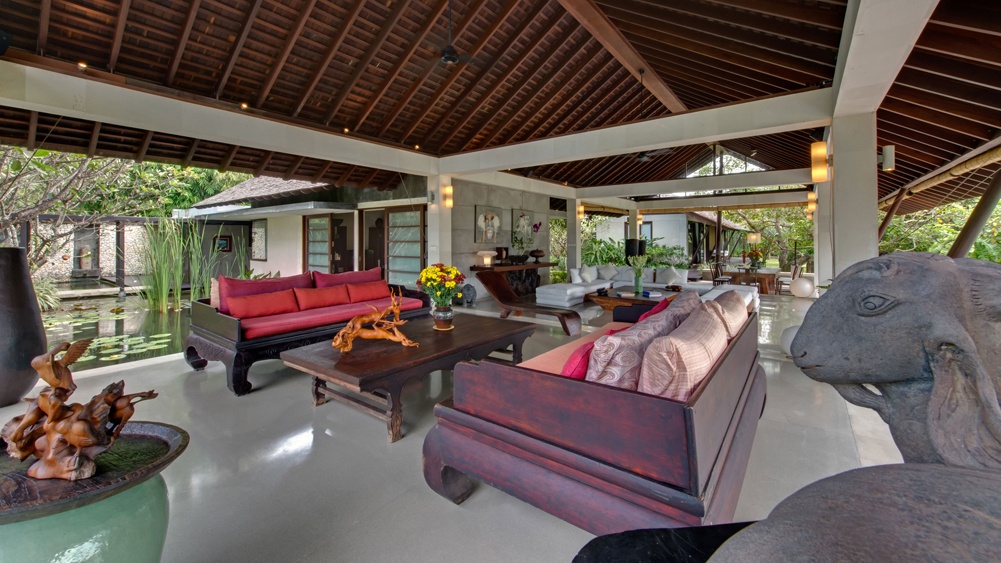 Villa Samadhana's open-air living pavilion beside a lotus pond, furnished with red and dark-wood daybeds under a vaulted roof