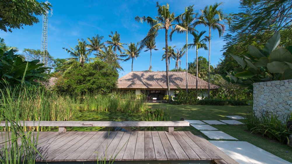 The gardens at Villa Samadhana with a lily pond and timber bridge, coconut palms and a thatched pavilion beyond