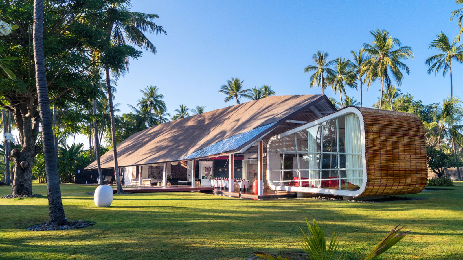 Long view across the lawn to the shingle-roofed main pavilion and its curved glass end at Villa Sapi, Lombok