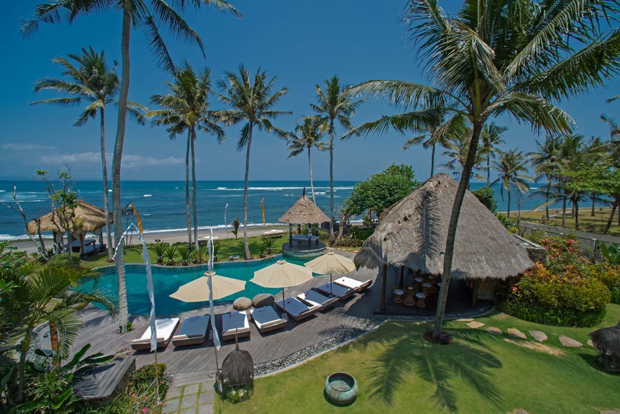 Aerial view of Villa Taman Ahimsa's beachfront pool, loungers, and thatched pavilions in Canggu, with the ocean behind