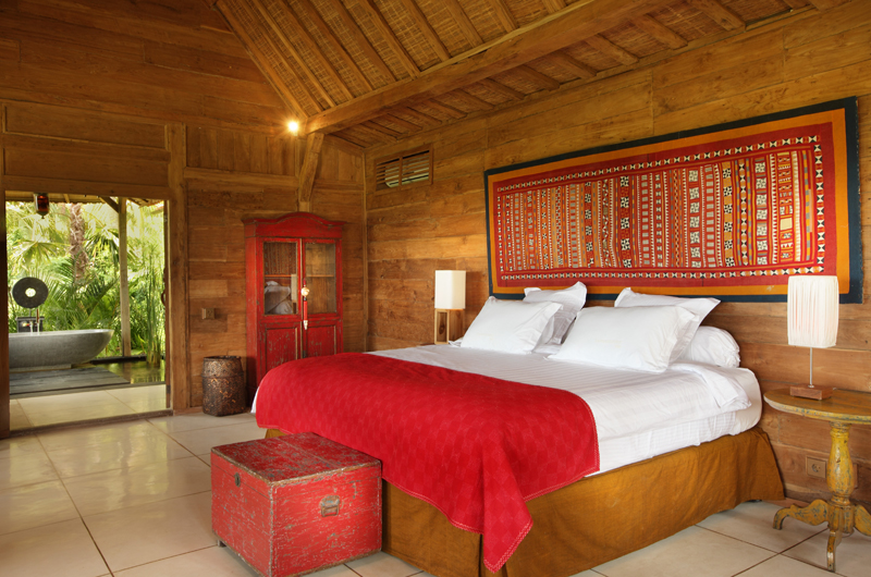 A teakwood bedroom at Villa Tangguntiti with a Balinese textile headboard and a stone garden bathtub beyond